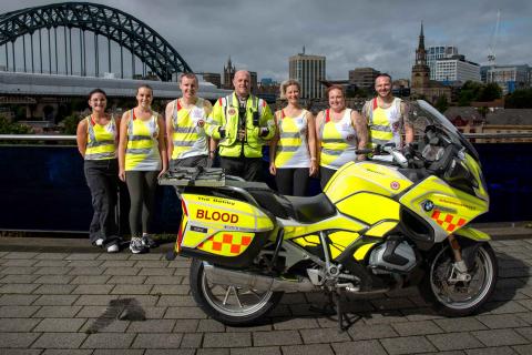 Group of team blood bike runners with a blood bike in front of the tyne bridge